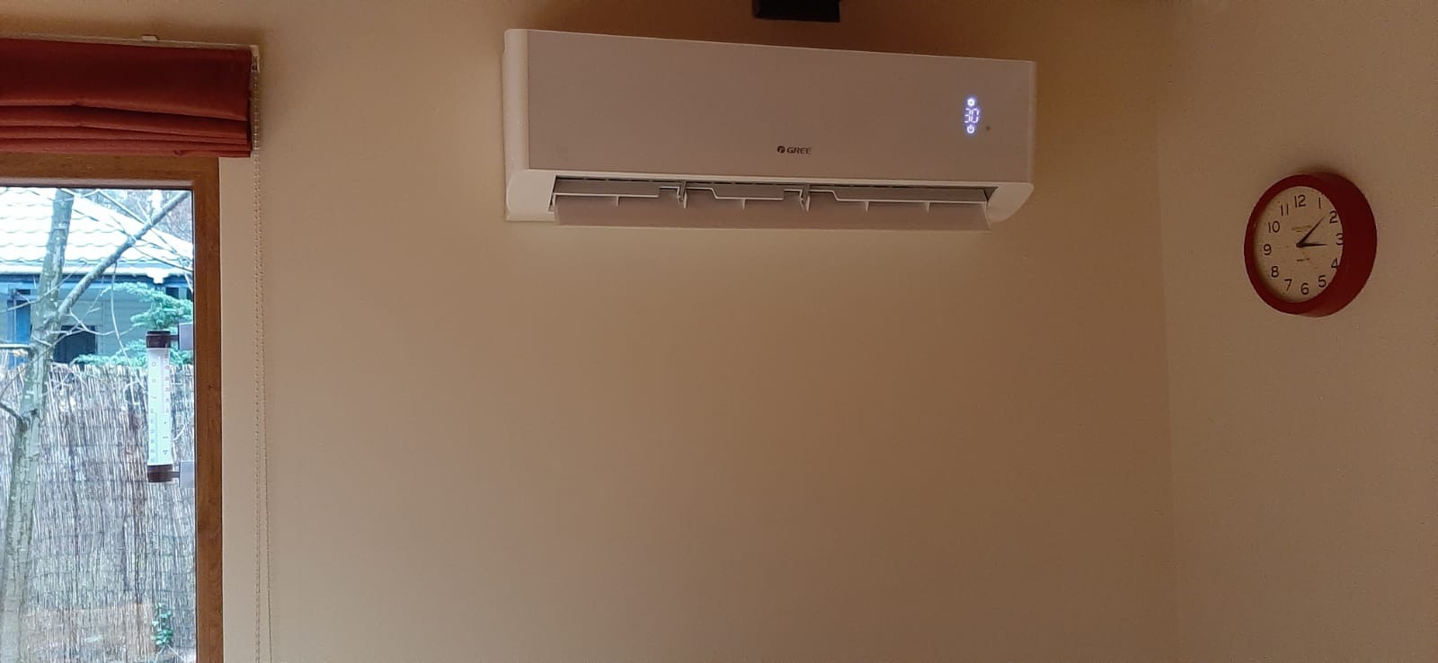 Biały klimatyzator marki Gree zamontowany na beżowej ścianie w pokoju, obok okna z roletą i czerwonego zegara wskazującego godzinę 14:10.
