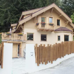 Dwukondygnacyjny dom z jasną elewacją, drewnianymi wykończeniami i balkonami z kwiatami, otoczony drewnianym ogrodzeniem z pionowych desek, na tle zielonego wzgórza i drzew.