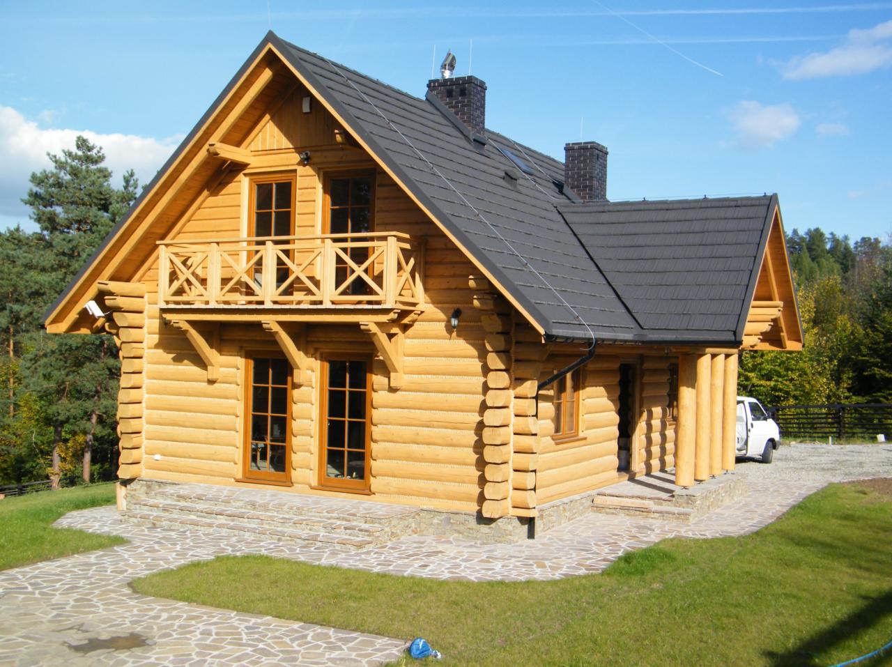 Nowoczesny dom z bali z drewnianym balkonem, kryty ciemnoszarą dachówką, z kamienną ścieżką i zadbanym trawnikiem.