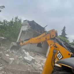 Żółta koparka JCB w trakcie rozbiórki budynku, widoczne gruzy i pracownik w tle.