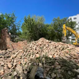 Żółta koparka Komatsu na tle błękitnego nieba i budynku mieszkalnego, usuwająca stertę gruzu ceglanego po rozbiórce, widoczne resztki muru i zieleń.
