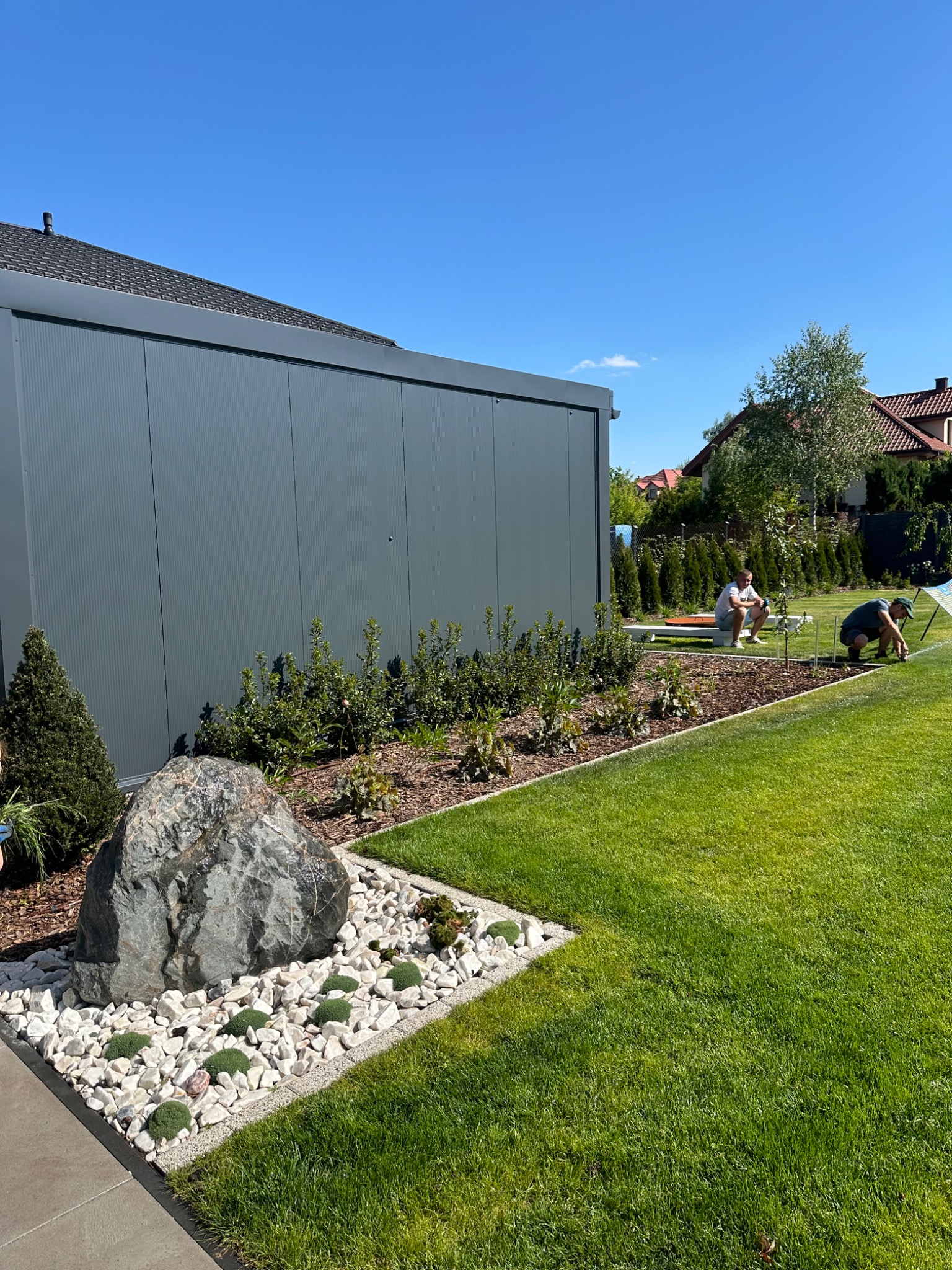 Aranżacja ogrodu z dużym kamieniem, białym żwirem i niskimi roślinami, w tle montaż systemu nawadniania przez dwóch mężczyzn, widok na trawnik i budynek gospodarczy.