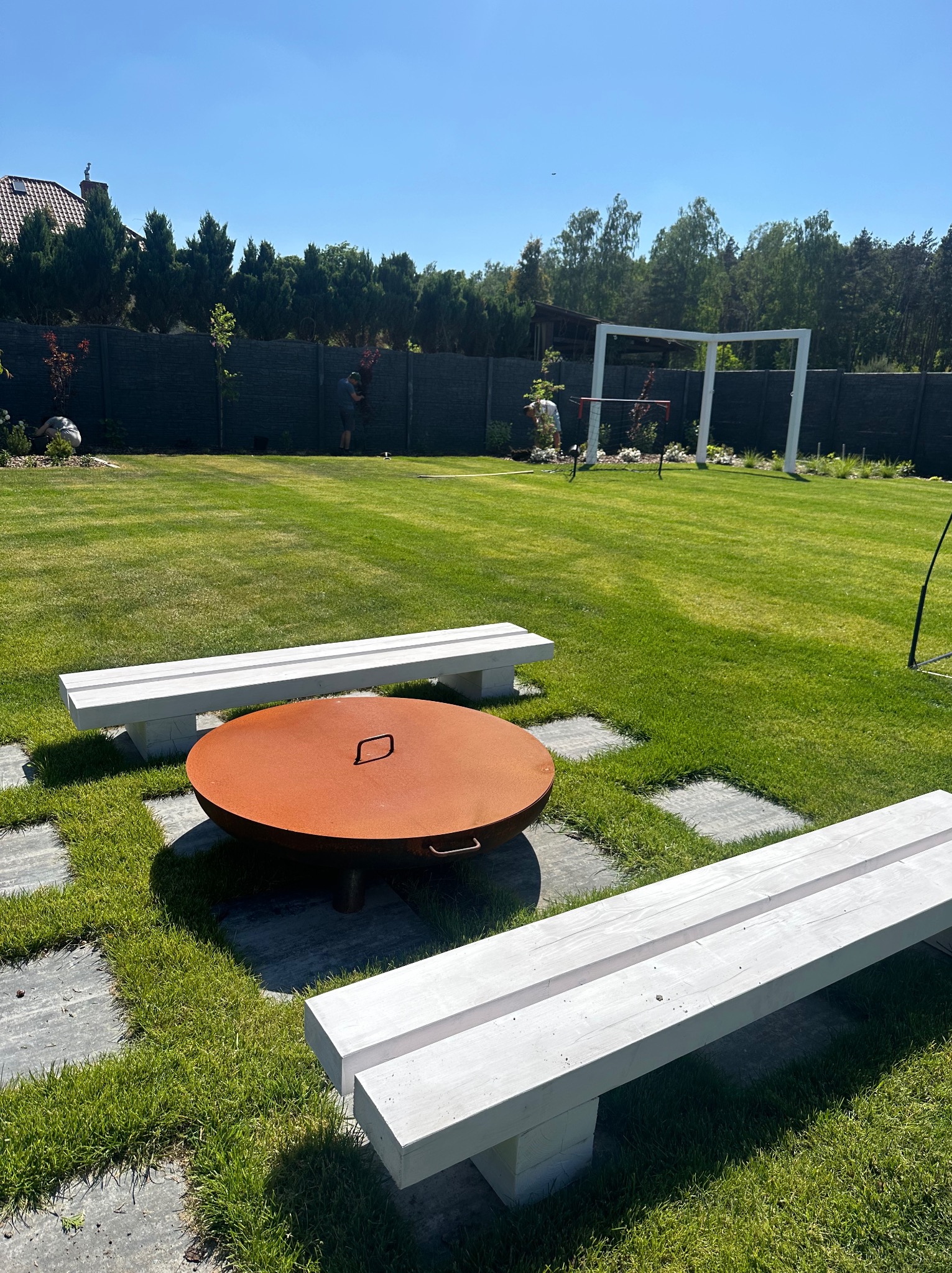 Przestronny ogród z równo przystrzyżonym trawnikiem, rustykalnym paleniskiem otoczonym betonowymi płytami i białymi ławkami, w tle osoby pracujące nad roślinnością wzdłuż ogrodzenia.