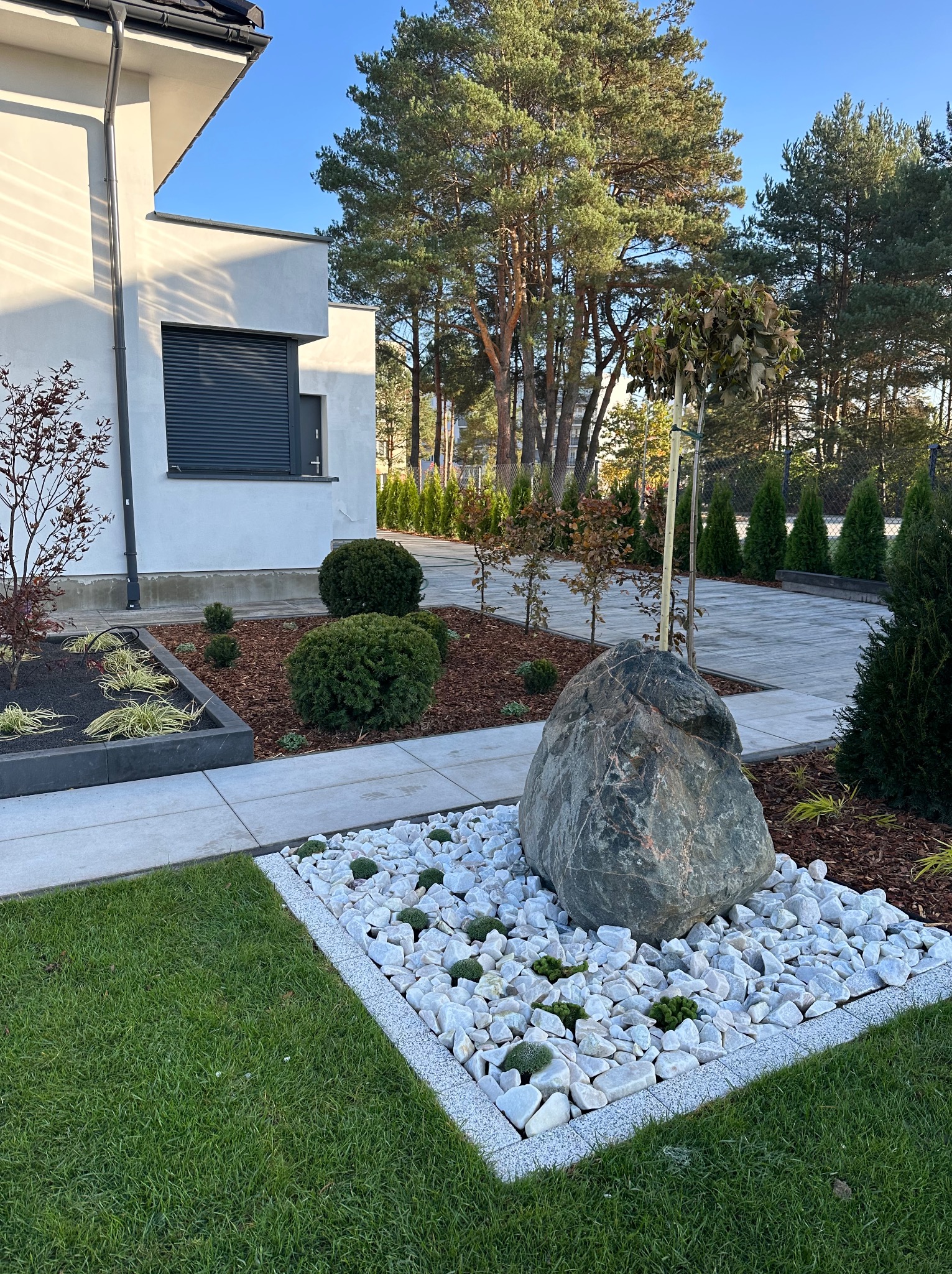 Nowoczesny ogród przydomowy z dużym, szarym kamieniem otoczonym białymi otoczakami i niską zielenią, z trawnikiem, rabatami z korą i roślinami, oraz ścieżką z płyt betonowych prowadzącą w głąb posesji