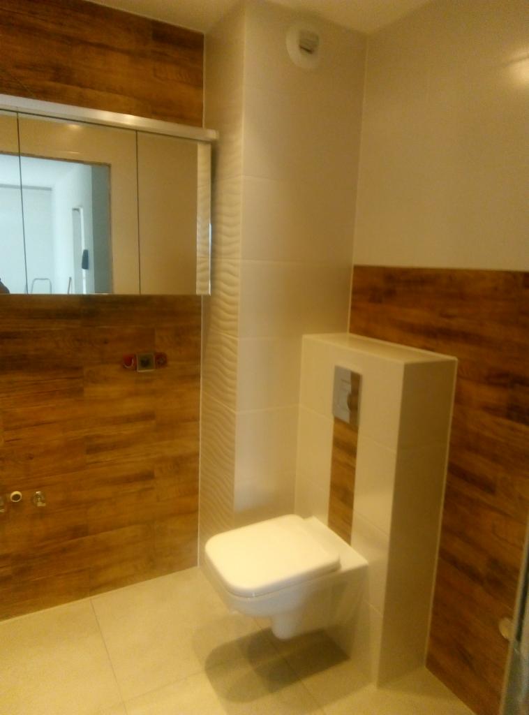 Nowoczesna toaleta podwieszana z zabudową, obłożona płytkami imitującymi drewno, na tle ściany z lustrem i dekoracyjnymi płytkami.