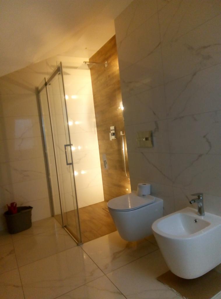 Nowoczesna łazienka z kabiną prysznicową ze szklanymi drzwiami, podwieszaną toaletą, bidetem i marmurowymi płytkami na ścianach i podłodze, oświetlona światłem zza ściany z drewnopodobnymi panelami.