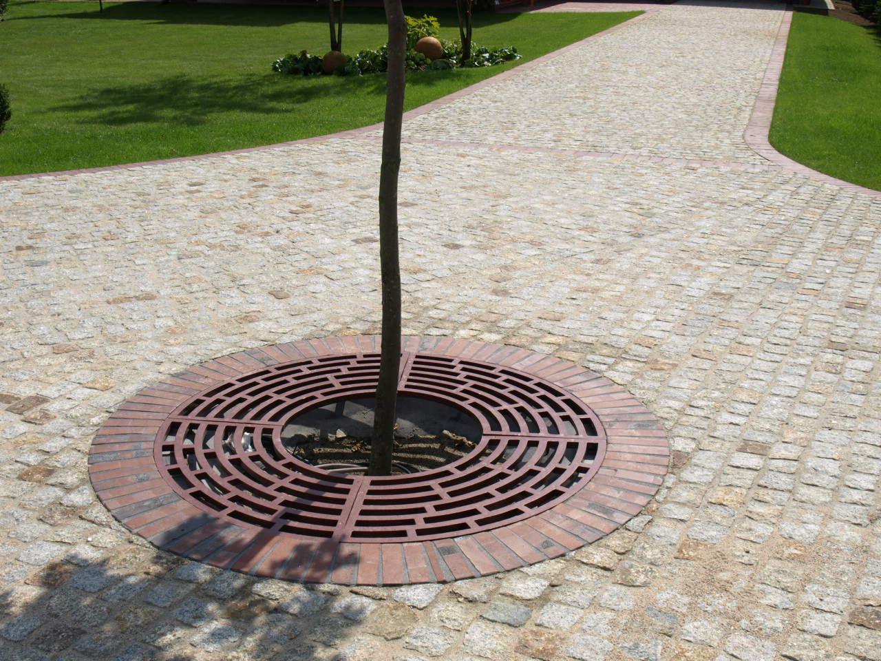 Brukowana ścieżka z granitu, otaczająca drzewo z dekoracyjną, okrągłą kratą w kolorze ceglastym, otoczoną obrzeżem z tego samego materiału. Trawa i drzewa w tle.