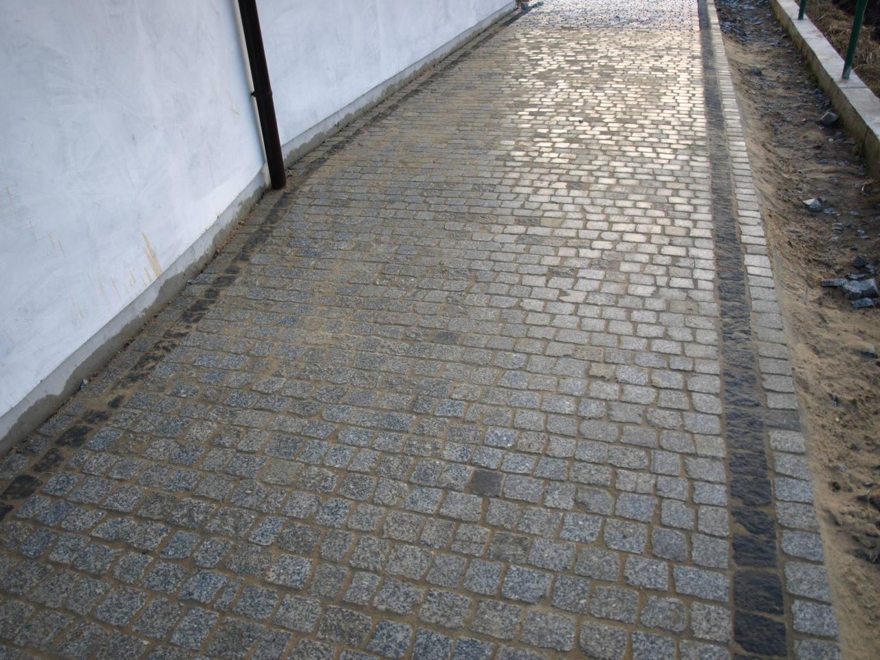 Ułożona kostka brukowa z szarego granitu tworząca ścieżkę, obok ściana budynku z rynną spustową i pas z ciemniejszej kostki oddzielający ścieżkę od gruntu.