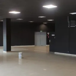 Wnętrze biura po malowaniu na ciemny kolor, widoczne dwa wiadra farby na błyszczącej podłodze i narzędzia malarskie w rogu, duże okna z widokiem na zewnątrz.