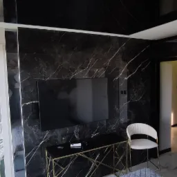 Eleganckie wnętrze z czarną marmurową ścianą, na której wisi telewizor. Złota konsola pod telewizorem, białe krzesło i regał w tle. Widok na balkon z białymi krzesłami.