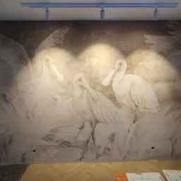 Ściana pokryta tapetą z motywem stada białych ptaków na beżowym tle, oświetlona listwą z reflektorami, z widocznymi przewodami elektrycznymi i fragmentem parkietu na podłodze.