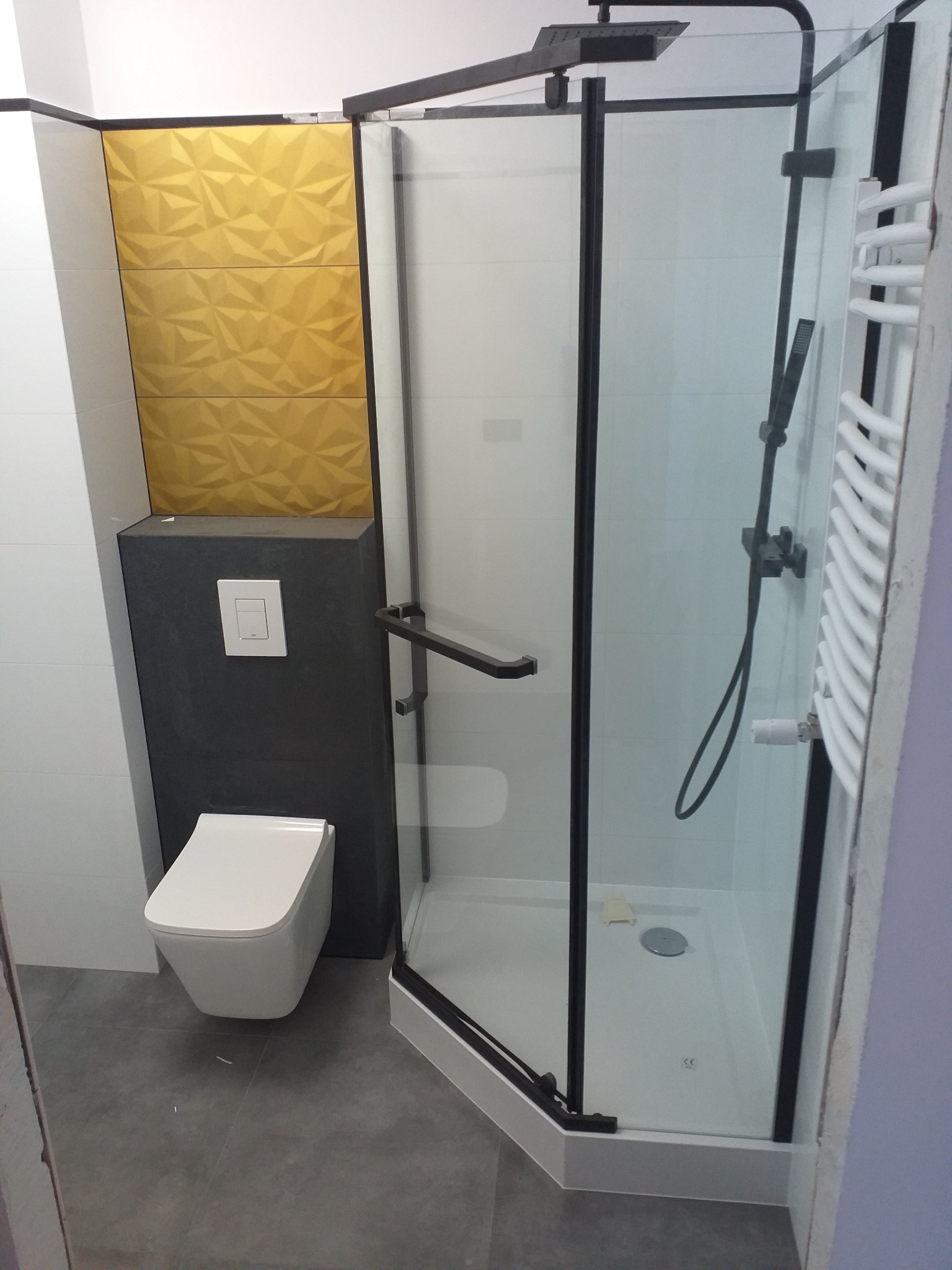 Nowoczesna łazienka z kabiną prysznicową z czarnymi ramami, wiszącą toaletą i złotymi płytkami 3D na ścianie, widok z boku.