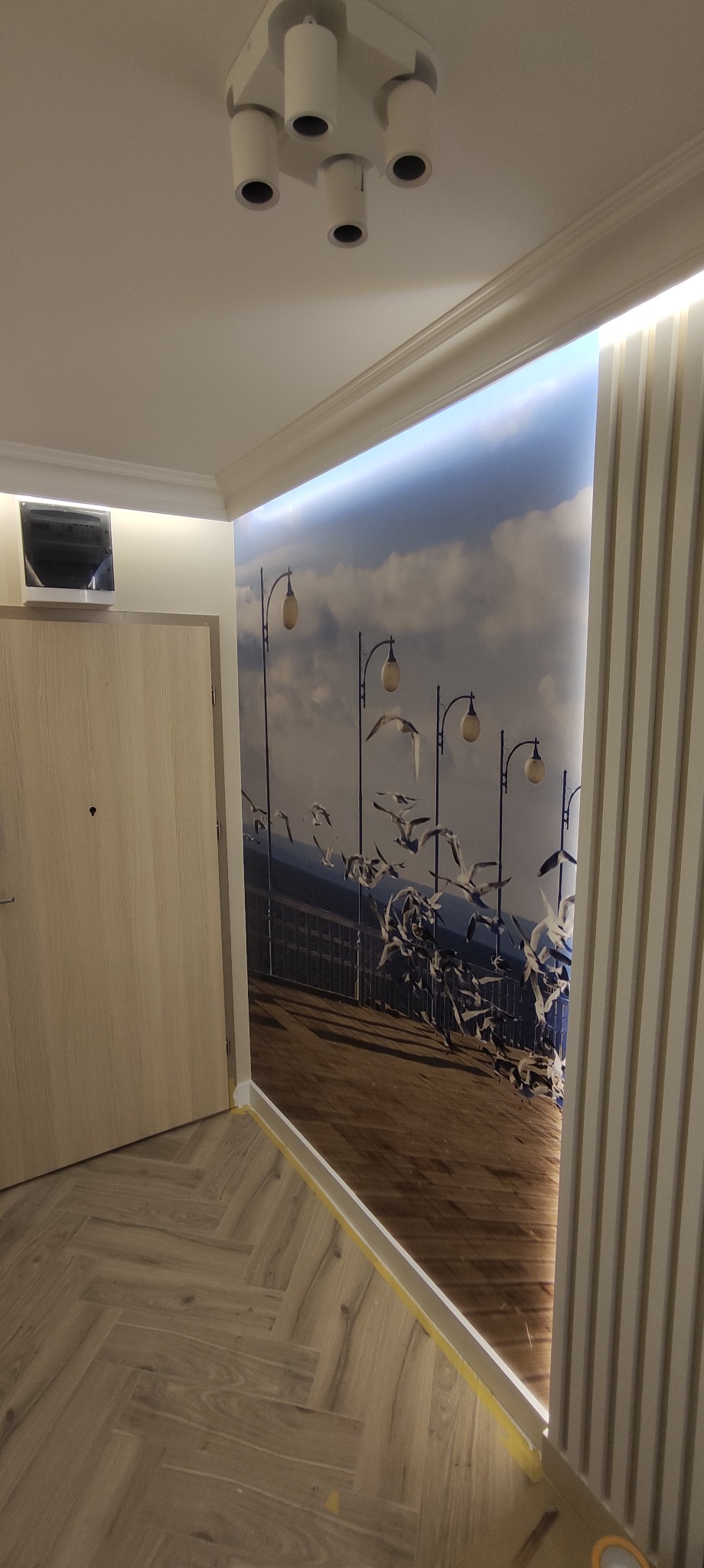 Fragment pokoju z fototapetą przedstawiającą molo i mewy, oświetlenie punktowe na suficie, drzwi w odcieniach beżu i panele ścienne w pionowe paski.