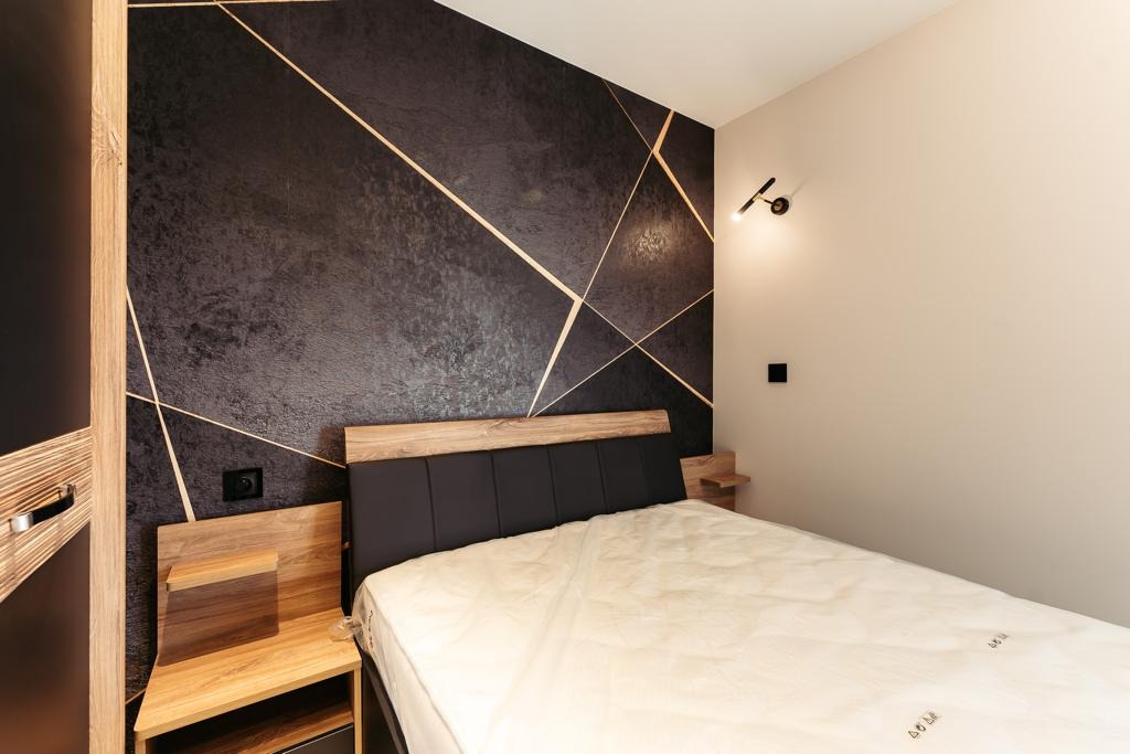 Czarna ściana w sypialni z abstrakcyjnym wzorem złotych linii, łóżko z ciemnym wezgłowiem i drewnianymi szafkami nocnymi, minimalistyczna lampka ścienna.
