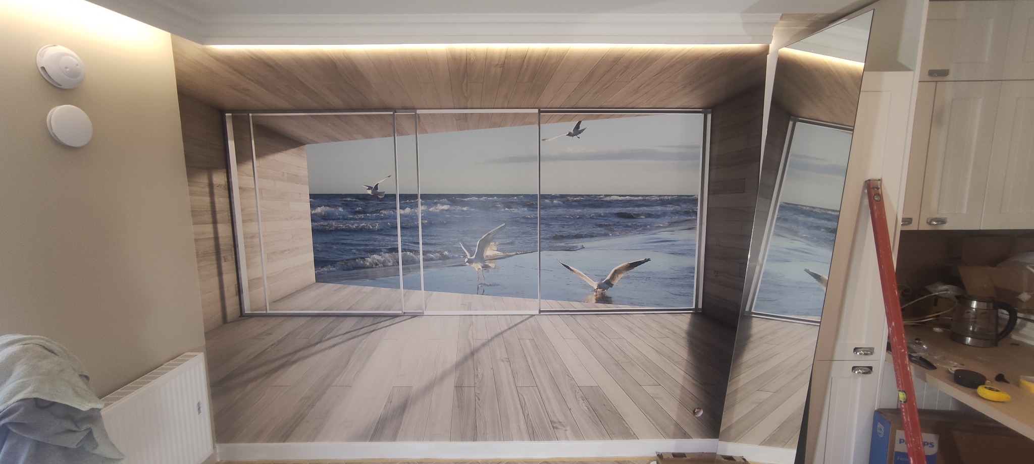 Fototapeta z motywem morza i mew na ścianie w pokoju, widoczne przesuwne panele fototapety, fragment ściany z grzejnikiem i poziomica oparta o ścianę.