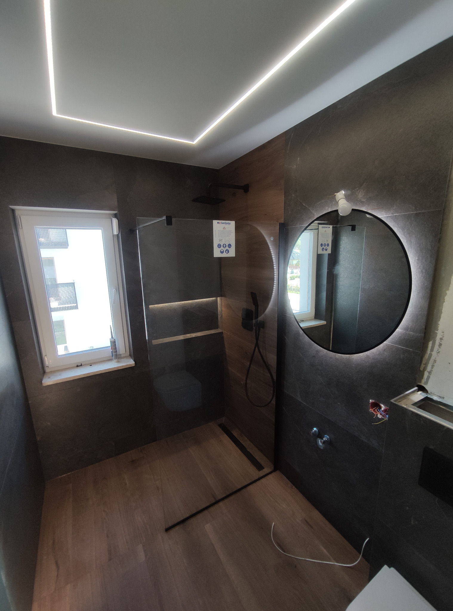 Nowoczesna łazienka z ciemnymi płytkami imitującymi kamień, drewnopodobną podłogą, kabiną prysznicową ze szklaną ścianką i oświetlonym lustrem okrągłym. Widoczne niedokończone prace przy instalacji...