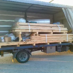 Ciężarówka z otwartą przestrzenią ładunkową, wypełnioną paletami z drewnem, kartonami i materiałami budowlanymi, zabezpieczonymi pasami.