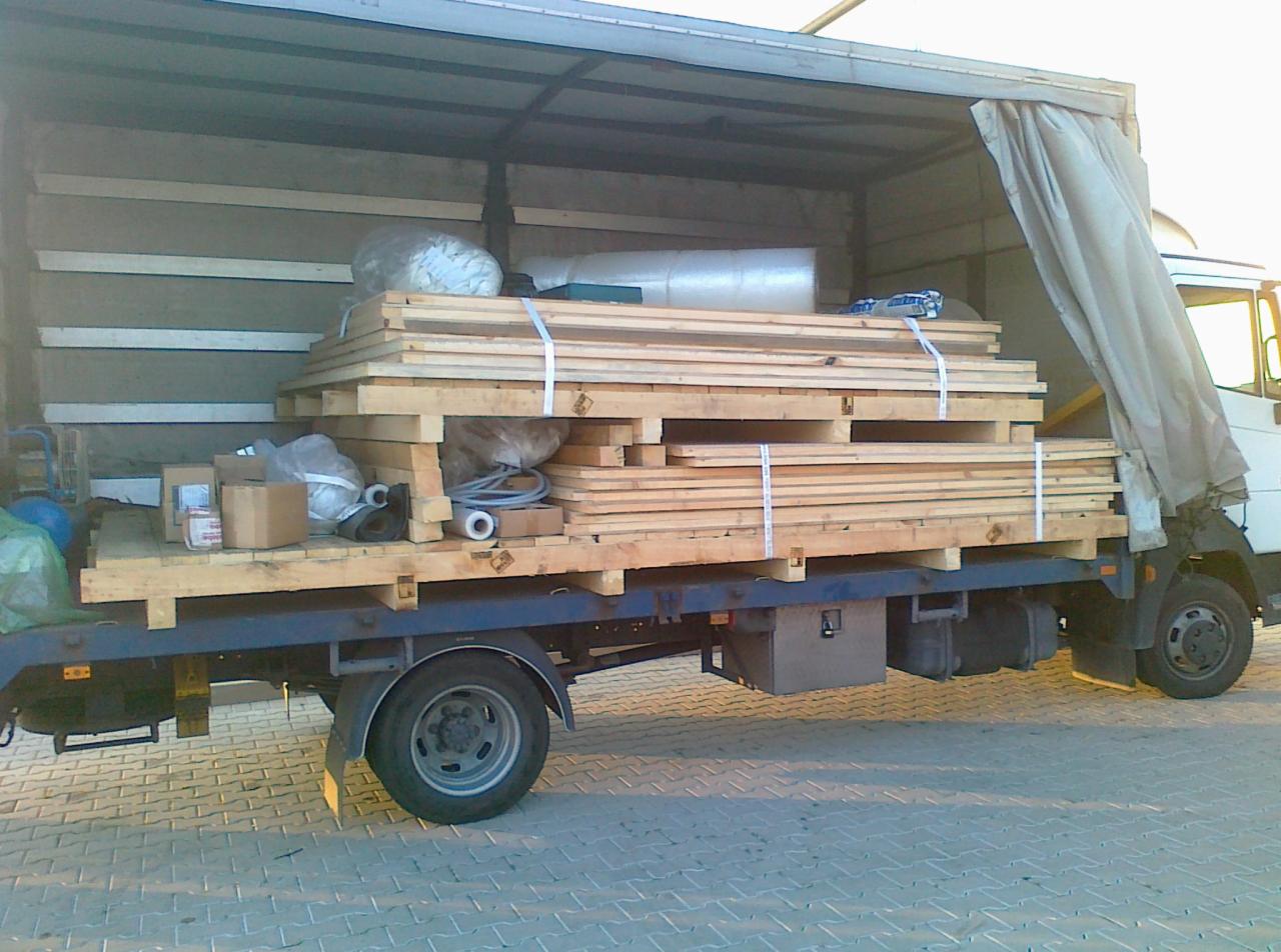 Ciężarówka z otwartą przestrzenią ładunkową, wypełnioną paletami z drewnem, kartonami i materiałami budowlanymi, zabezpieczonymi pasami.