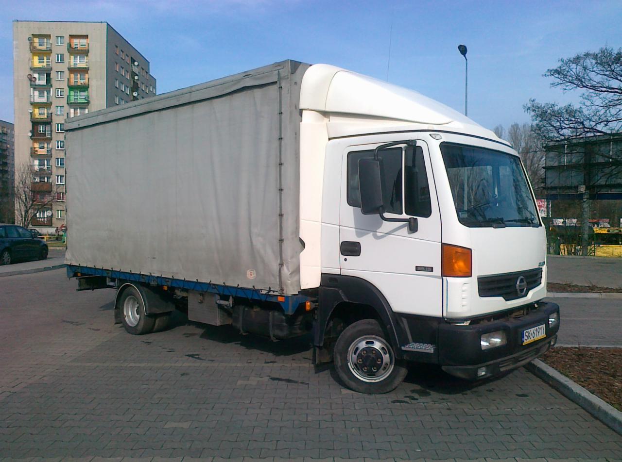 Biały samochód dostawczy z plandeką na tle bloku mieszkalnego. Widoczny numer rejestracyjny SK 6191Y. Dzienny kadr z chodnika.