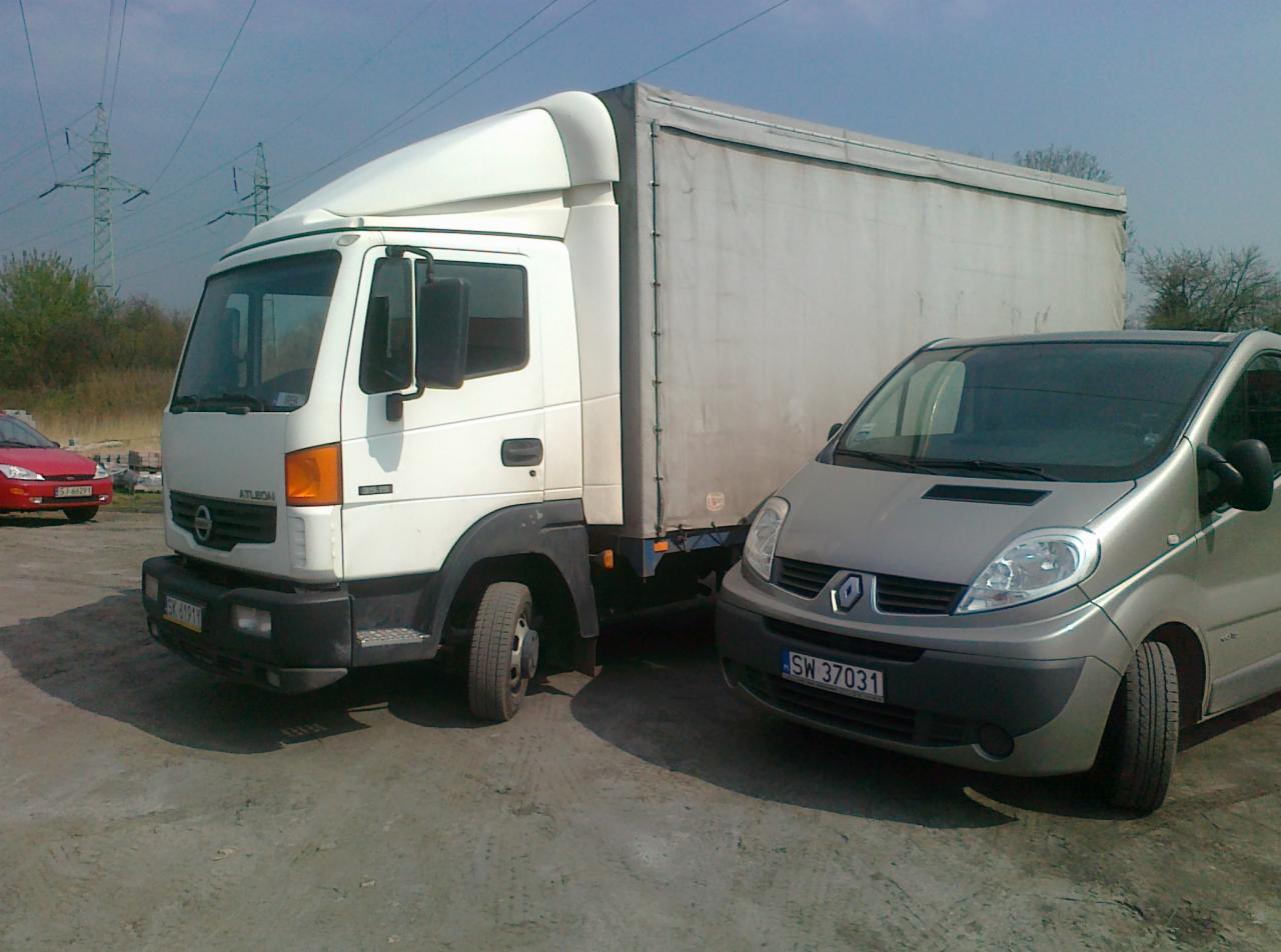Biały samochód dostawczy Nissan Atleon z plandeką i srebrny Renault Trafic na placu, w tle słupy wysokiego napięcia.