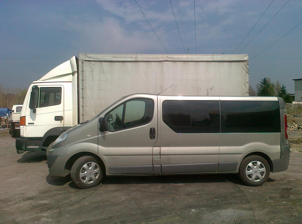 Srebrny bus Renault Trafic z przyciemnianymi szybami, zaparkowany obok białej ciężarówki z plandeką na szarym placu.