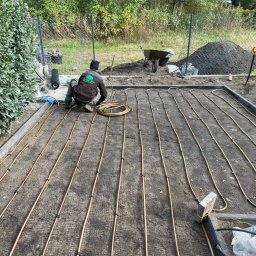 Garden Art Katarzyna Klimkowska - Instalacja systemu nawadniania pod trawnik: pracownik układa rury na siatce, widoczny kopiec ziemi i narzędzia ogrodnicze. Etap montażu przed zasypaniem.