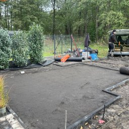 Garden Art Katarzyna Klimkowska - Przygotowany teren pod trawnik z rolki, obrzeża ułożone, ziemia wyrównana, pracownik przy samochodzie z rolkami trawy w tle, rośliny w donicach z boku.