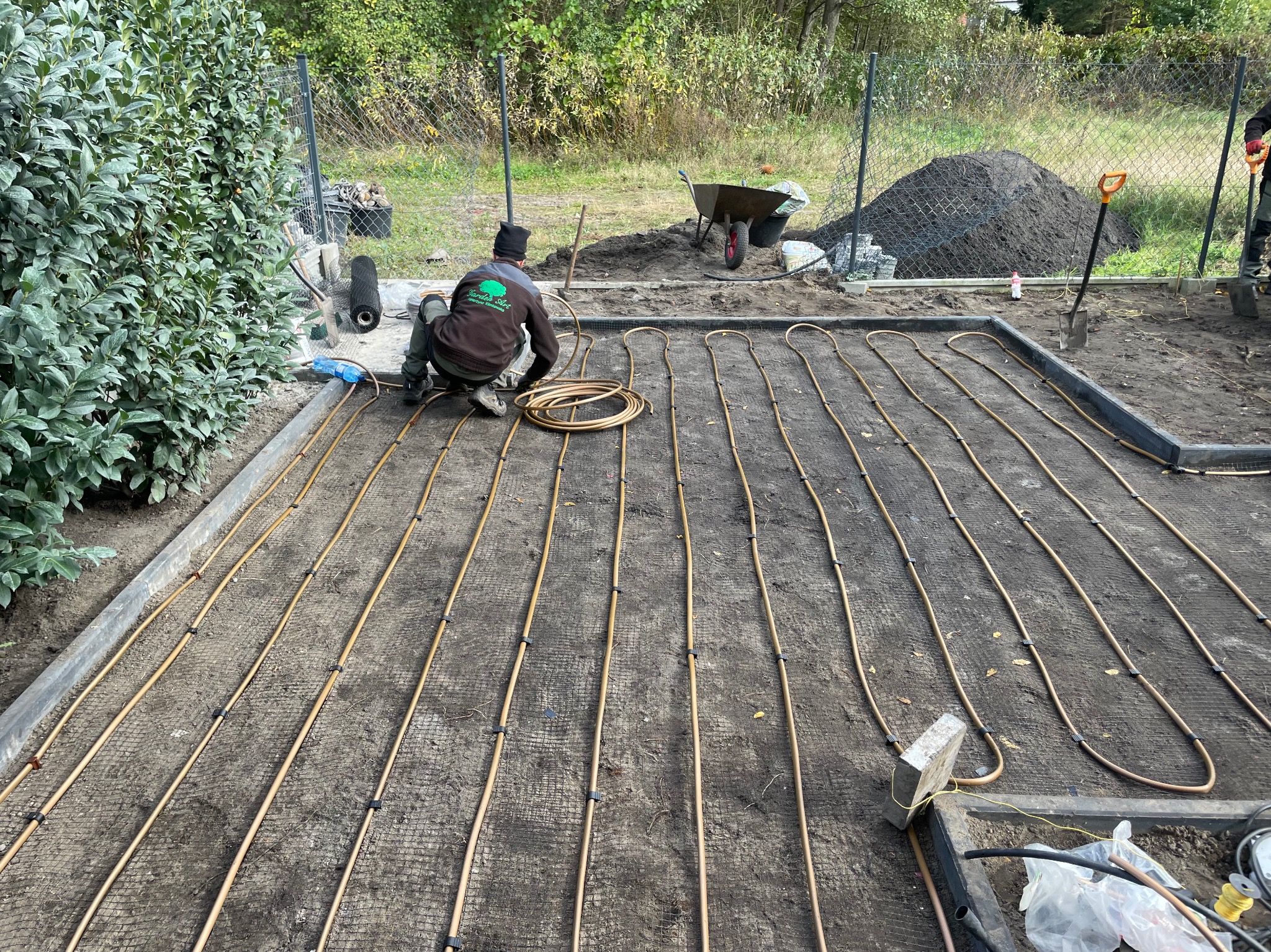Instalacja systemu nawadniania pod trawnik: pracownik układa rury na siatce, widoczny kopiec ziemi i narzędzia ogrodnicze. Etap montażu przed zasypaniem.