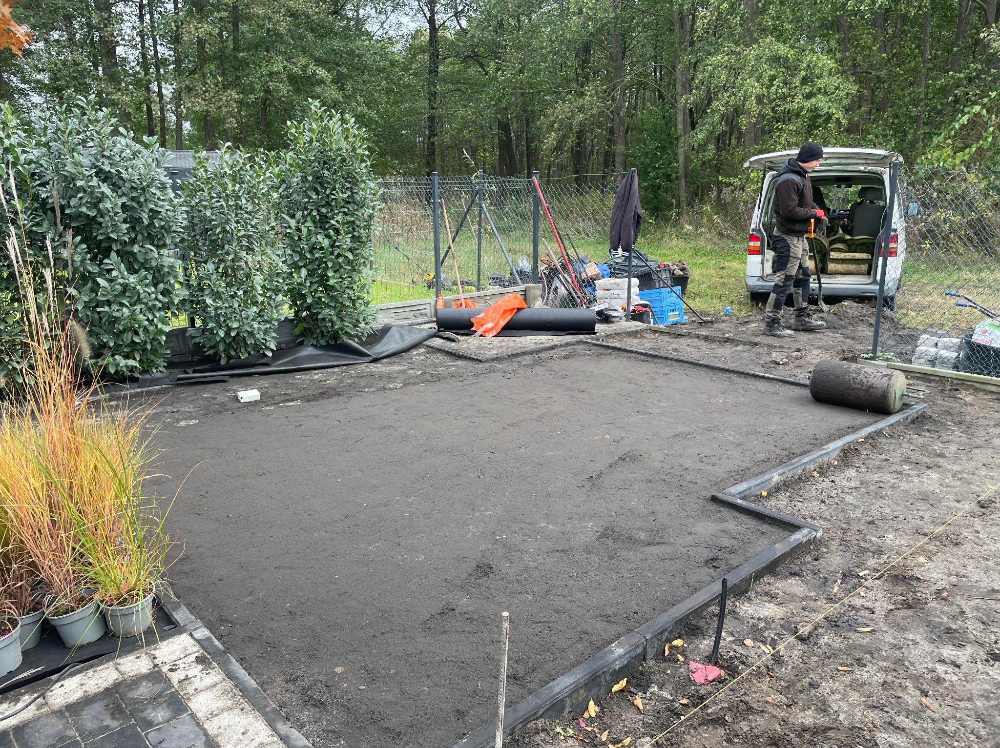 Przygotowany teren pod trawnik z rolki, obrzeża ułożone, widoczny pracownik i narzędzia. W tle drzewa i samochód dostawczy.