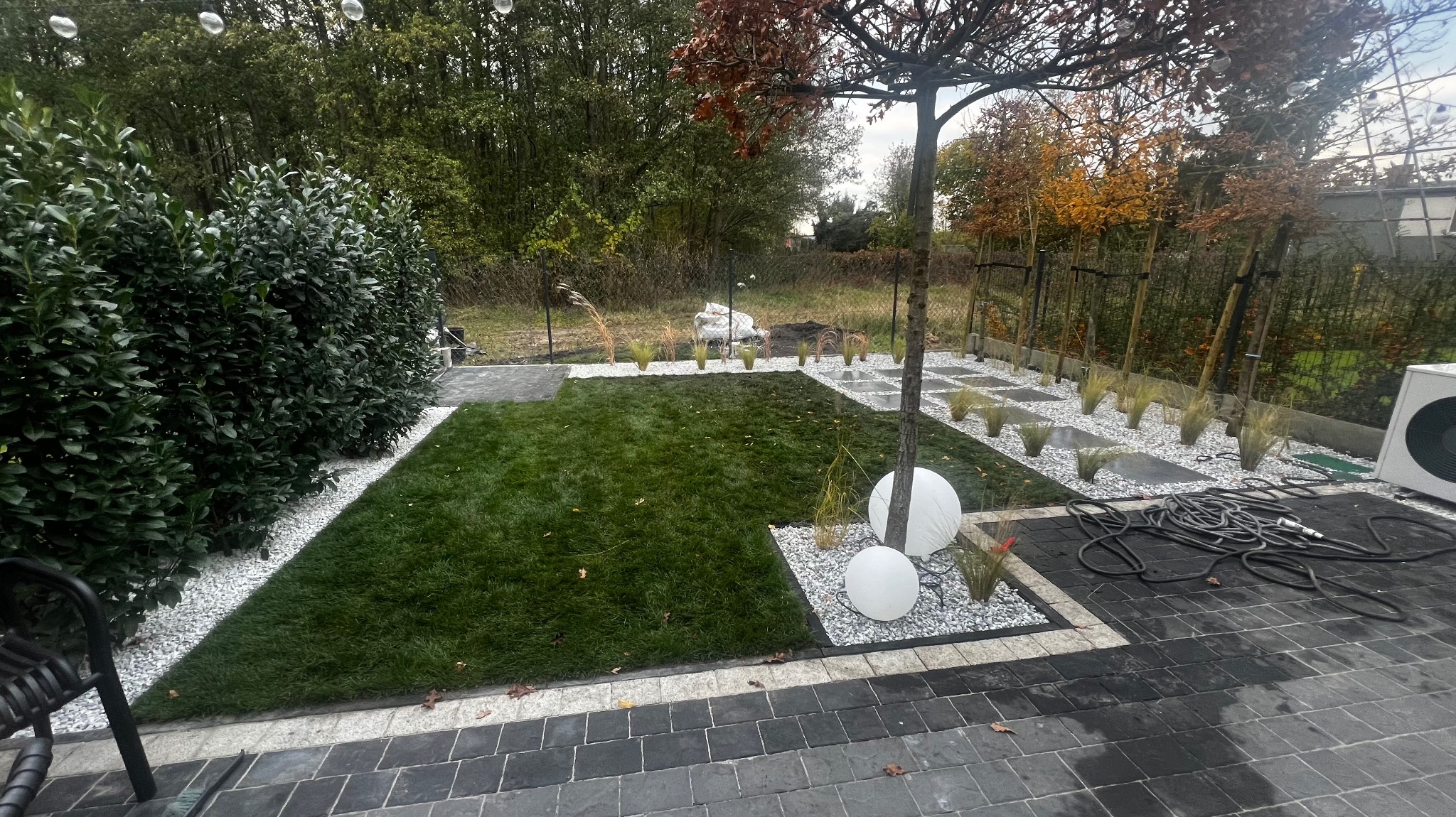 Nowoczesny ogród z trawnikiem, białymi kamieniami i kulistymi lampami. Widoczne świeżo posadzone rośliny i starannie ułożona kostka brukowa. Elementy systemu nawadniania.