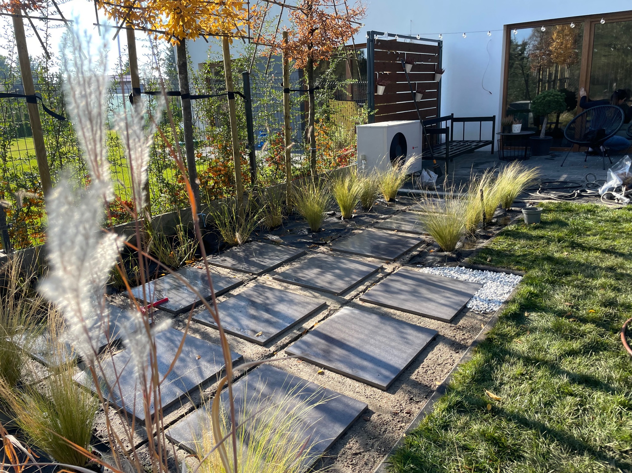 Układanie ścieżki z płyt w ogrodzie z ozdobnymi trawami i białym żwirem. Widoczny fragment trawnika i ogrodzenie. W tle budynek z dużymi oknami.