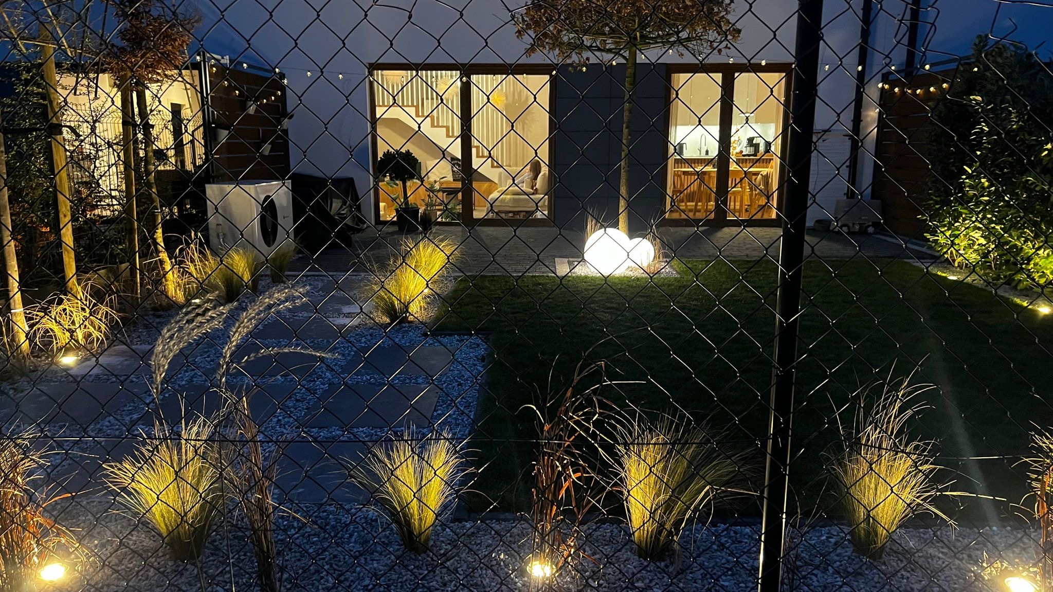 Oświetlony ogród nocą, widok przez siatkę ogrodzeniową. Dekoracyjne trawy, ścieżka z kamyków, podświetlane kule i budynek w tle z oknami.
