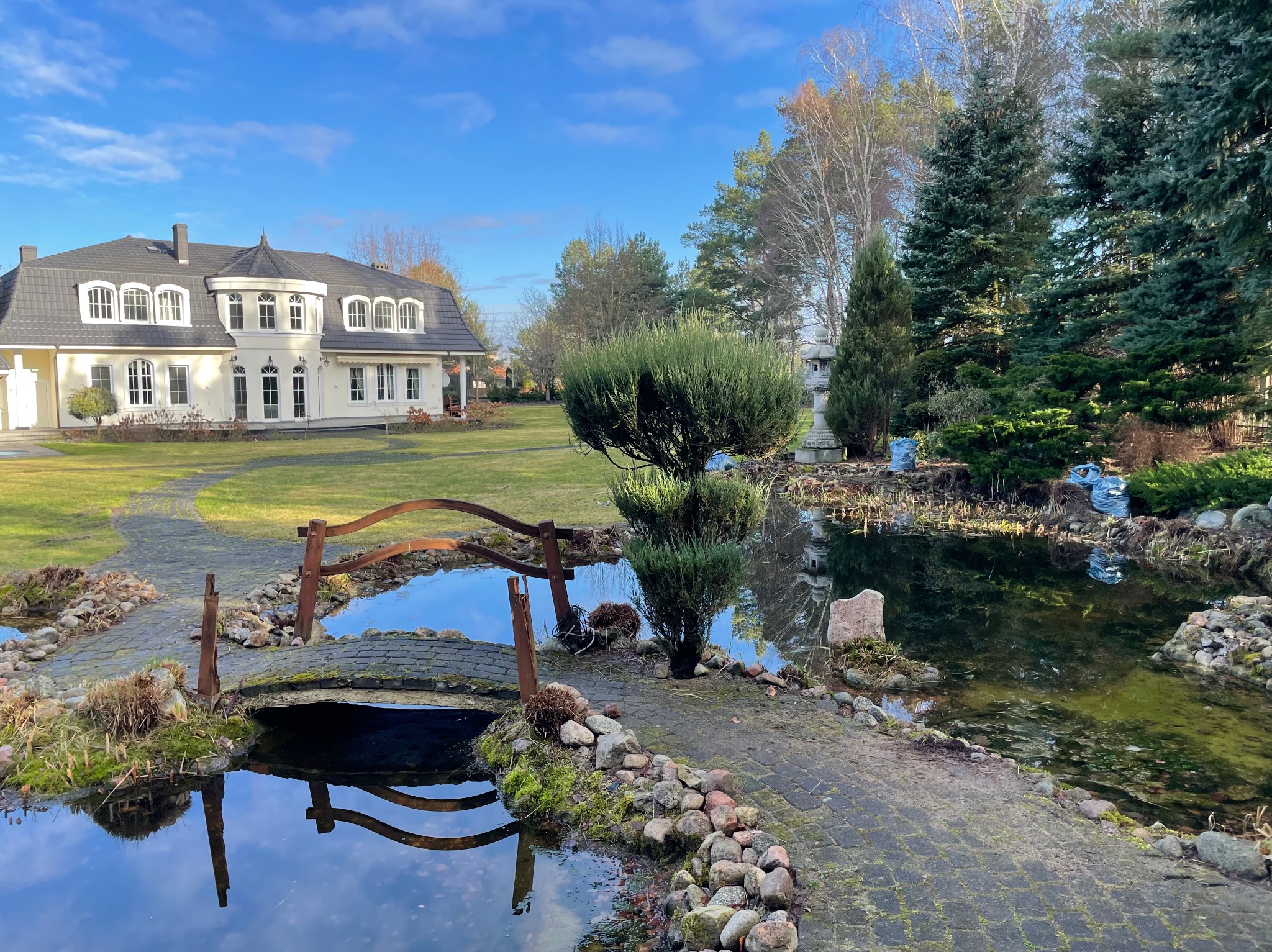 Ogród z małym mostkiem nad stawem i eleganckim domem w tle. Starannie zaprojektowana przestrzeń z elementami wodnymi i roślinnością.