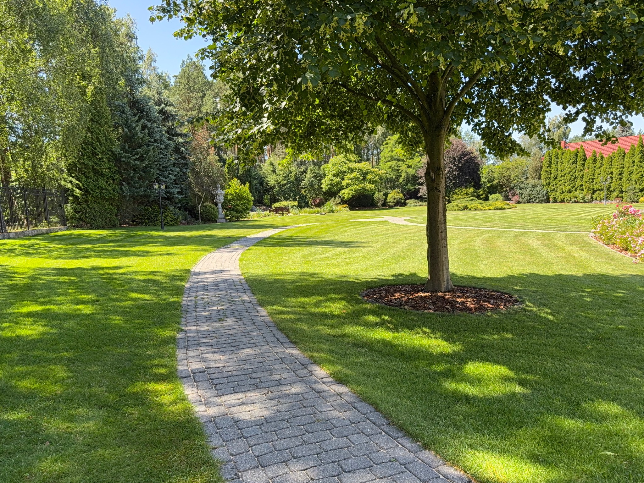 Zadbany ogród z trawnikiem w paski, ścieżką z kostki brukowej i drzewami. Widoczne krzewy i kwiaty. Oświetlenie słoneczne, podkreślające zieleń trawy.