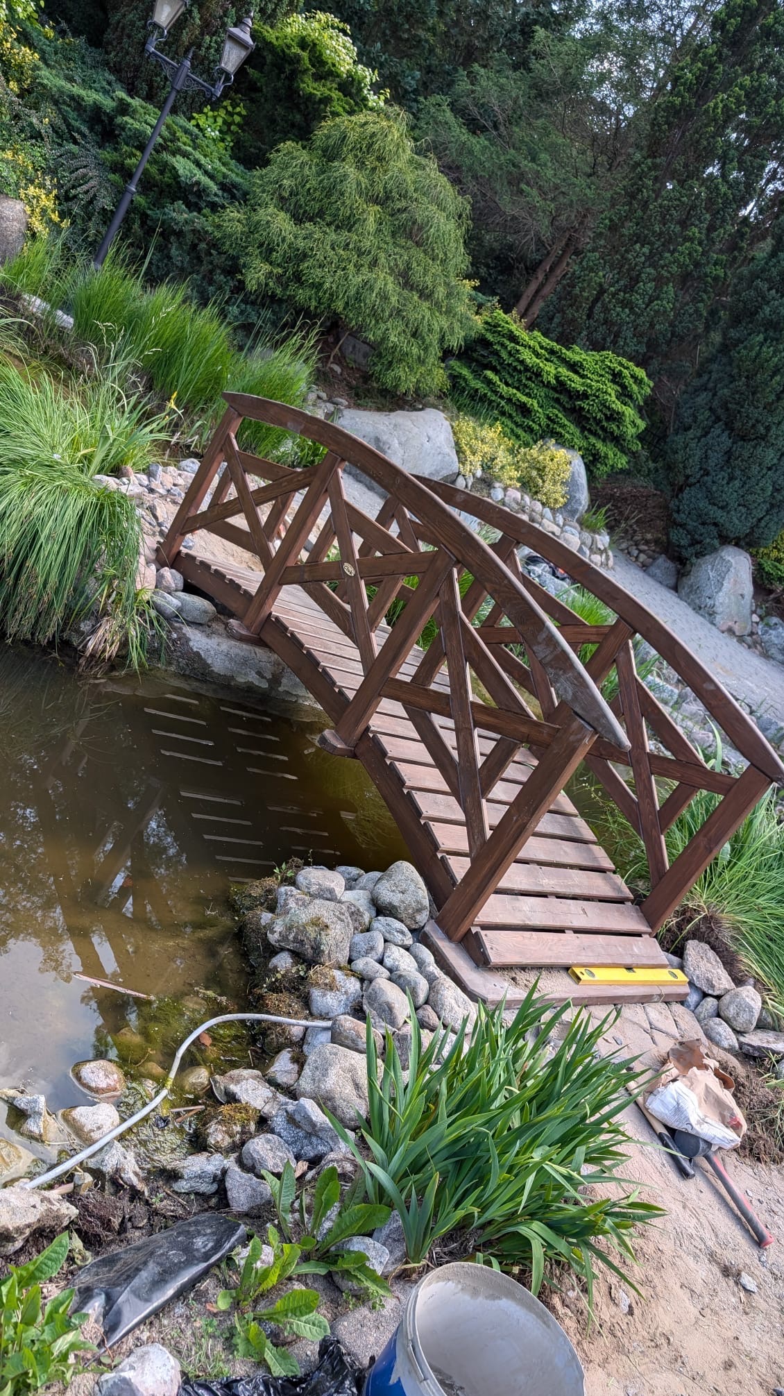 Drewniany mostek nad oczkiem wodnym w ogrodzie, otoczony kamieniami i roślinnością. Widoczne narzędzia i materiały budowlane sugerują prace wykończeniowe.
