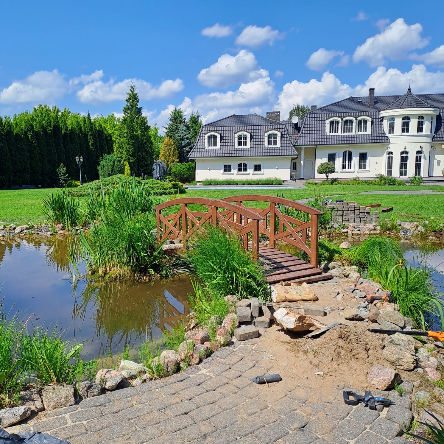 Ogród z oczkiem wodnym i drewnianym mostkiem w trakcie budowy. Widoczne narzędzia i materiały budowlane. W tle duży dom i błękitne niebo z chmurami.