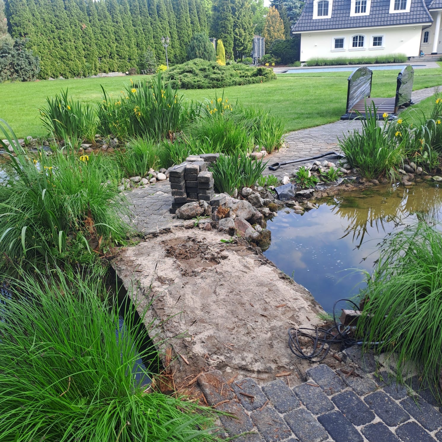 Budowa mostku nad oczkiem wodnym w ogrodzie: ułożone kamienie, trawa, w tle dom z basenem. Prace w toku, widoczne narzędzia i materiały budowlane.