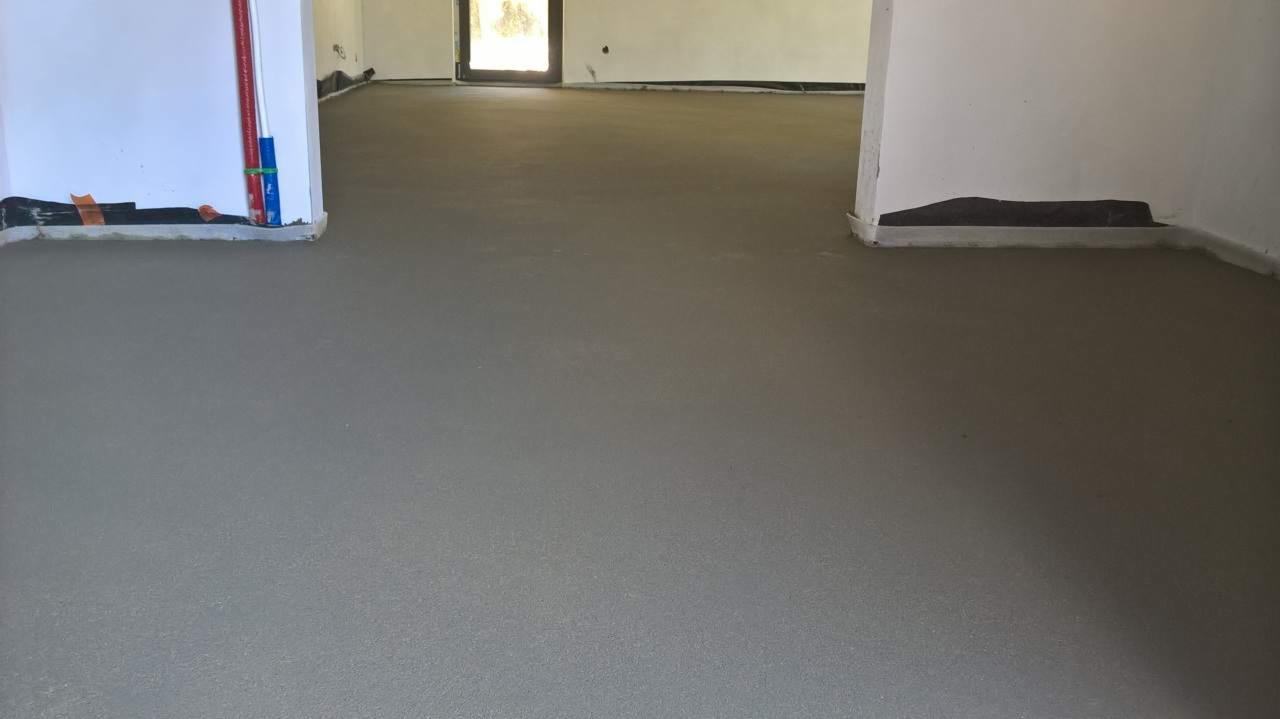 Świeżo wylana, gładka, szara posadzka w surowym wnętrzu z widocznymi instalacjami przy ścianach i zabezpieczeniami na dole ścian.