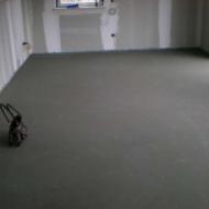 Świeżo wylana, szara posadzka w surowym pomieszczeniu z widocznymi śladami prac wykończeniowych na ścianach i niewielkim oknem w tle. Na podłodze leżą narzędzia.