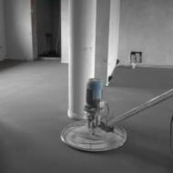 Szlifowanie wylewki samopoziomującej maszyną z długim ramieniem w pomieszczeniu z betonowym słupem i otwartym przejściem do innego pokoju.