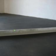 Wyrównywanie ciemnej, jednolitej powierzchni podłogi długą poziomicą z libellą w centralnej części kadru, przy białej ścianie.