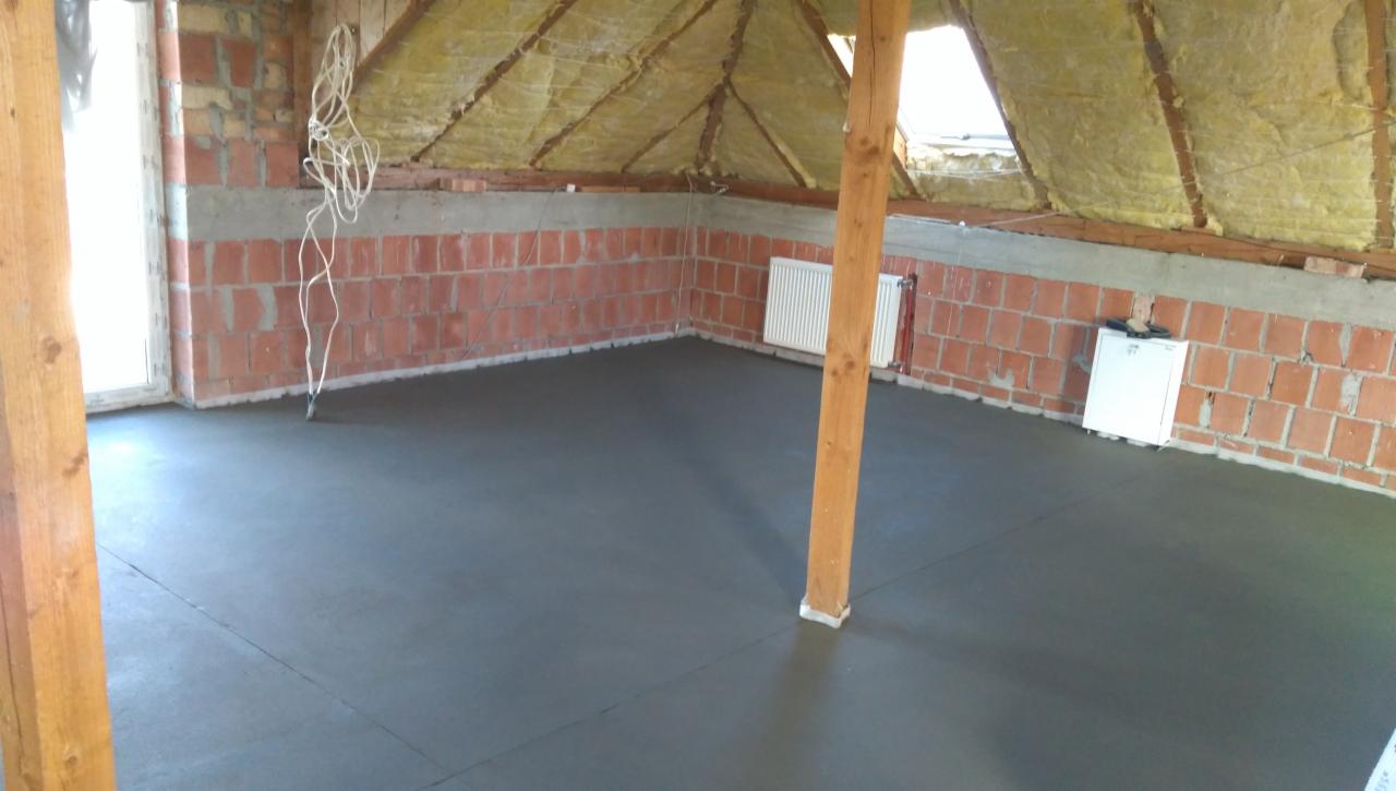 Szara, świeżo wylana posadzka w surowym pomieszczeniu na poddaszu z ceglanymi ścianami, drewnianym słupem i ocieplonym dachem, widoczne kable i grzejnik.