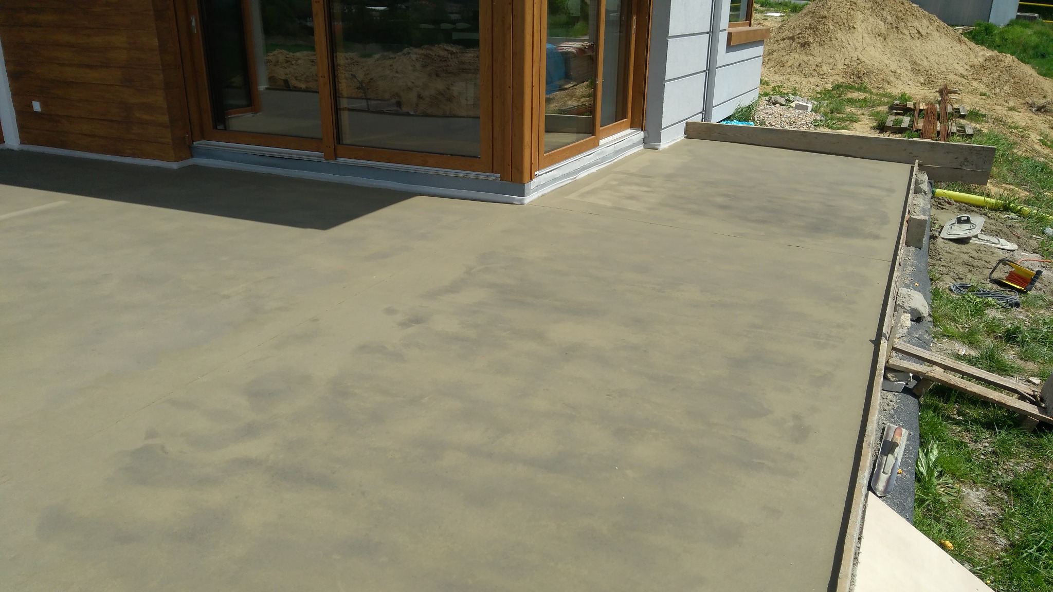 Świeżo wylana, gładka posadzka na zewnątrz budynku, widoczne drewniane szalunki i narzędzia budowlane, w tle okna z drewnianymi ramami i hałda piasku.