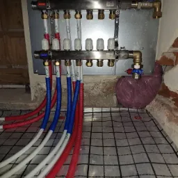 Rozdzielacz instalacji grzewczej na ścianie, z podłączonymi rurami w kolorach czerwonym i niebieskim, ułożonymi na folii z nadrukowaną siatką, fragment ściany z cegły w tle.