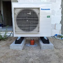 Biała jednostka zewnętrzna klimatyzacji Panasonic R32 zamontowana na betonowych bloczkach z gumowymi podkładkami, odpływ skroplin skierowany w dół, widoczne przewody i ściana budynku w stanie surowym.