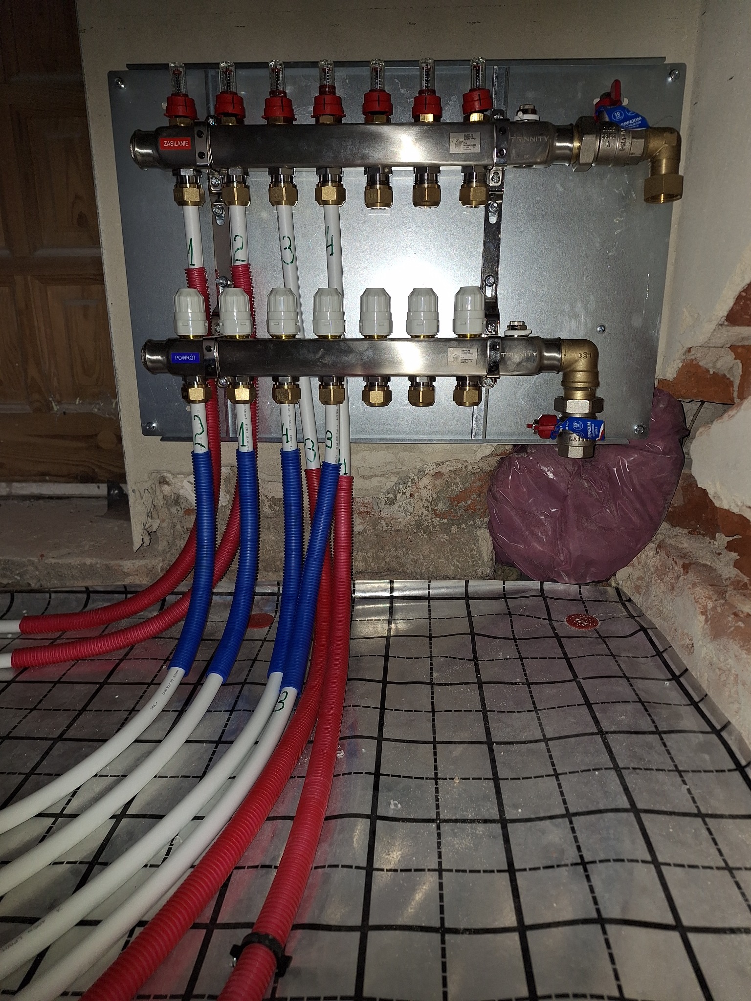 Rozdzielacz instalacji grzewczej na ścianie, z podłączonymi rurami w kolorach czerwonym i niebieskim, ułożonymi na folii z nadrukowaną siatką, fragment ściany z cegły w tle.