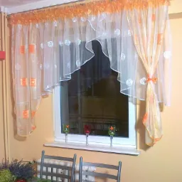 Okno z firankami w odcieniach bieli i pomarańczy, z motywem kwiatowym, upięte kokardą, na parapecie trzy kieliszki z kwiatami, w tle stół z zielonym obrusem.