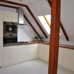 Jasna kuchnia na poddaszu z drewnianymi belkami, w zabudowie z wbudowanym ekspresem do kawy i piekarnikiem. Blat drewniany, białe fronty, płytki podłogowe.