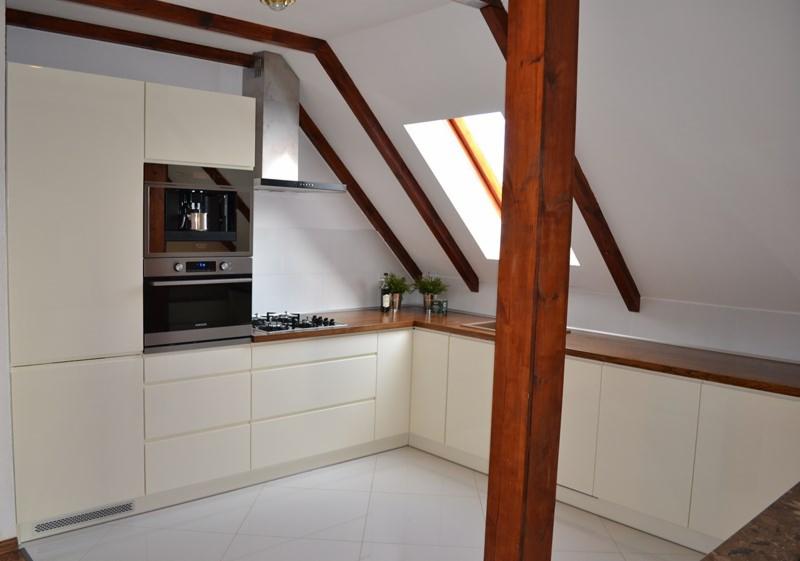 Jasna kuchnia na poddaszu z drewnianymi belkami, w zabudowie z wbudowanym ekspresem do kawy i piekarnikiem. Blat drewniany, białe fronty, płytki podłogowe.