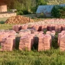 Ułożone w stosy, owinięte folią paczki z drewnem kominkowym na trawiastym terenie, z widocznym stosem nieopakowanego drewna w tle oraz budynkami w oddali.